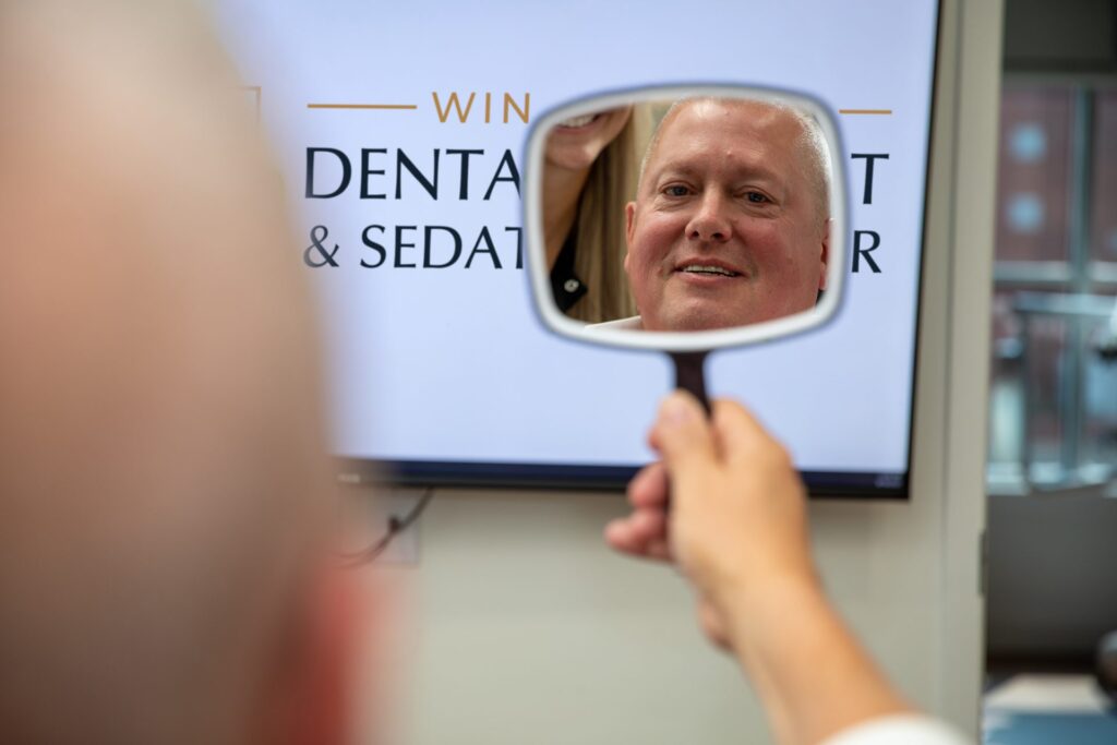 Upper All-on-4 patient smiling into a hand mirror in front of the Winchester Dental Implant & Sedation Center logo